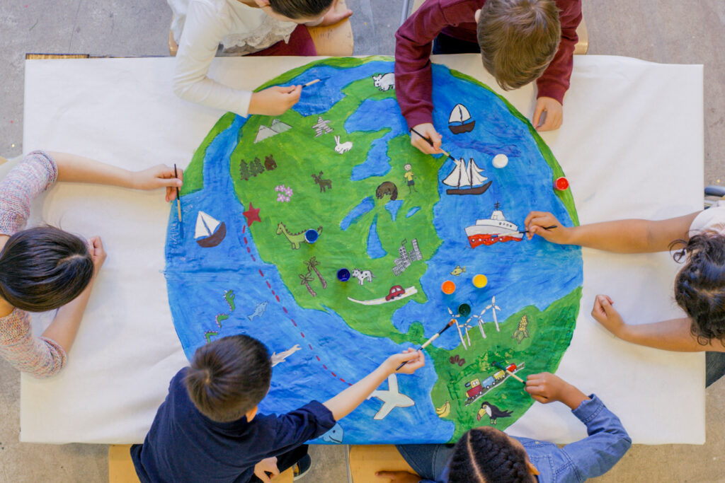 An aerial view of a multi ethnic group of children painting the globe and it's animals, fish, and vehicles. One girl paints a steam boat, while a boy colours in the ocean.
